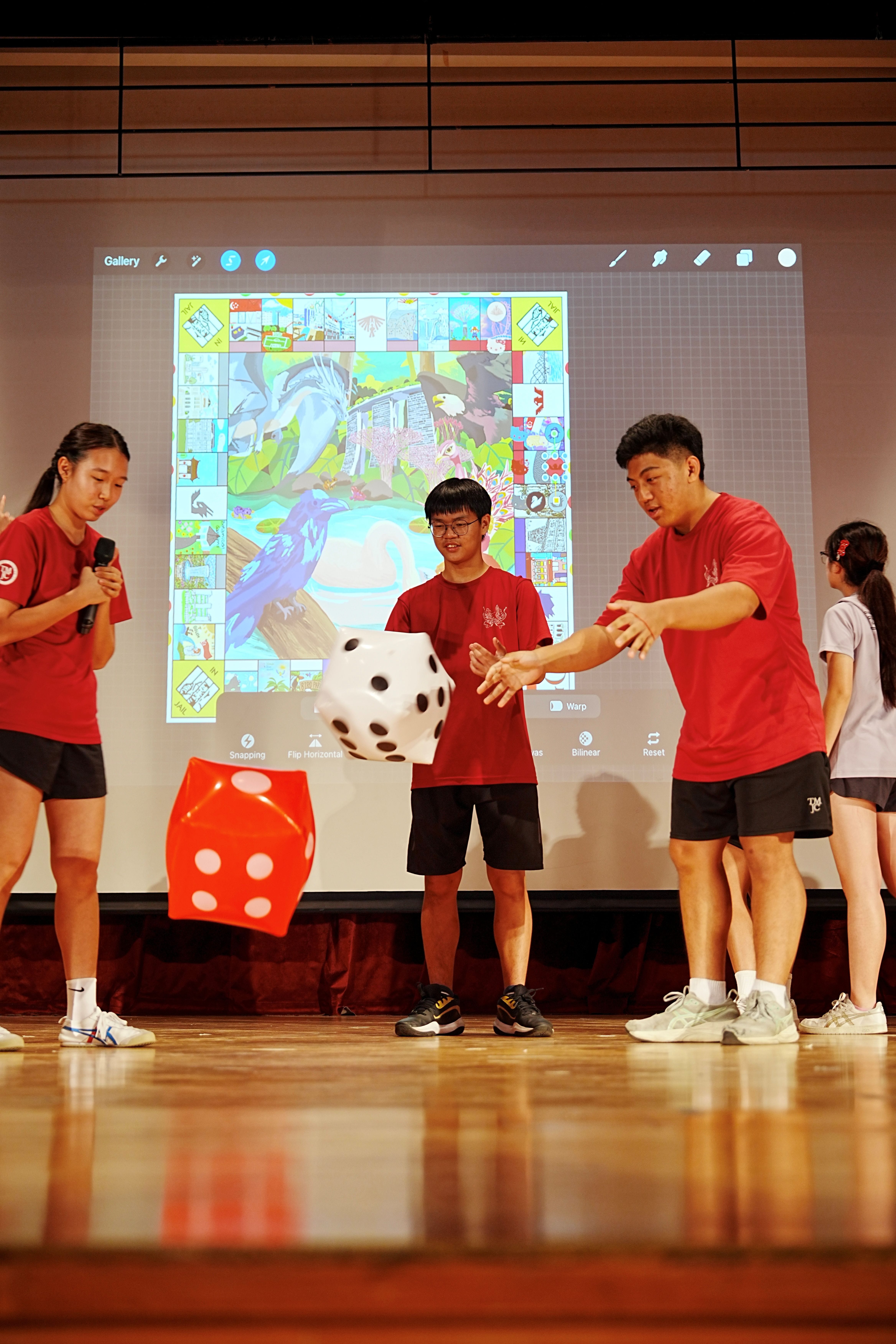 Three students in red T-shirts toss oversized dice on a stage in front of a projected screen displaying a vibrant, student-illustrated Monopoly-style board game called "TM-opoly."